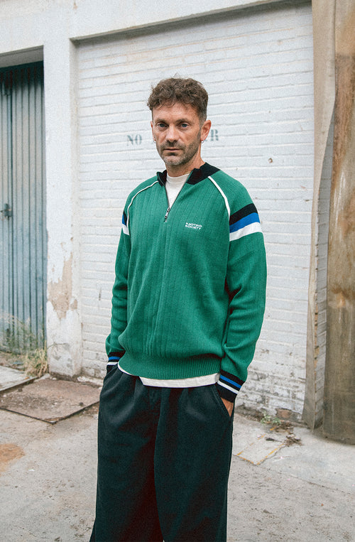 Jersey de punto verde con media cremallera, cuello alto y detalles deportivos en blanco, negro y azul.